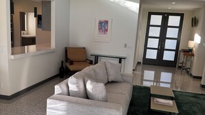 Uma sala de estar ampla com uma janela grande, um sofá, uma cadeira de madeira, uma mesinha e uma porta com painéis de vidro jateado.