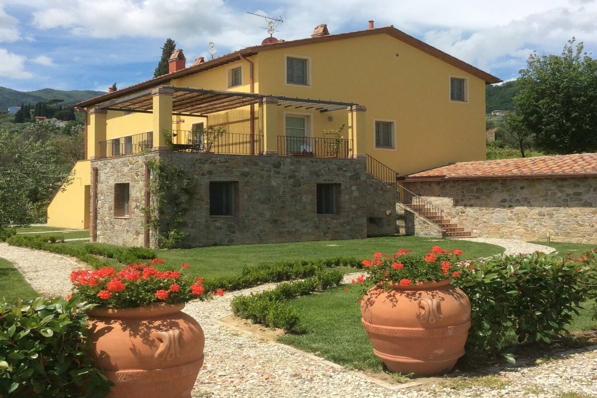 A yellow building with a stone base, a balcony, and a garden with potted plants.