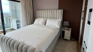 Habitación de hotel con una cama grande, una mesita de noche, una lámpara y vistas a la ciudad a través de un ventanal amplio.