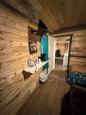 A wooden room with a bed, a small table with items, a door, and a wall-mounted TV.