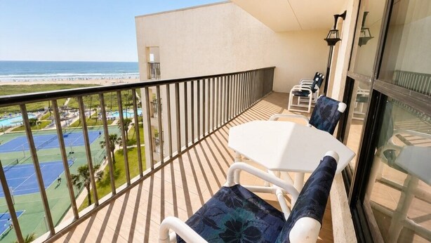 A balcony with a white table and chairs, overlooking a beach and tennis courts. - Saida IV Condos - S4904 (South Padre Island)
