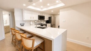 A modern kitchen with white cabinetry, a central island, and a breakfast bar with three wicker chairs. - Saida IV Condos - S4904 (South Padre Island)