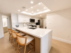A modern kitchen with white cabinetry, a central island, and a breakfast bar with three wicker chairs. - Saida IV Condos - S4904 (South Padre Island)