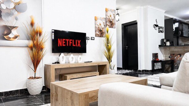 Ein modernes Wohnzimmer mit einem Flachbildfernseher, der 'NETFLIX' anzeigt, einem Holztisch, einer Couch und einer Küchenzeile im Hintergrund.