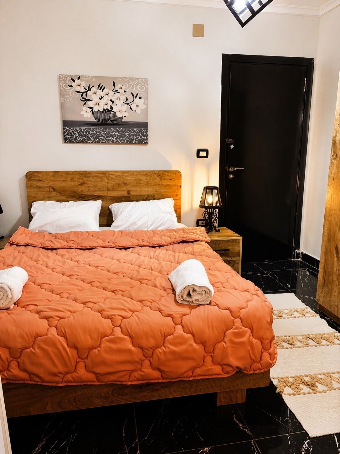 Ein ordentlich bezogenes Bett mit einem hölzernen Kopfteil, weißen Kissen und einer orangefarbenen Steppdecke. Darüber hängt ein Blumenbild, und zur rechten Seite ist eine schwarze Tür zu sehen.