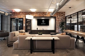 A modern living room with a large sectional sofa, a flat-screen TV mounted on a brick wall, and a long wooden table. - Arcadia Oasis w/ Resort Pool, Gym & Fire Pits (Phoenix)