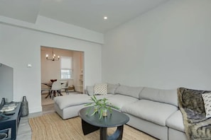 Ein modernes Wohnzimmer mit einem Ecksofa, einem kleinen runden Tisch mit Pflanzen, einem Essbereich mit Stühlen und einer Küche im Hintergrund.