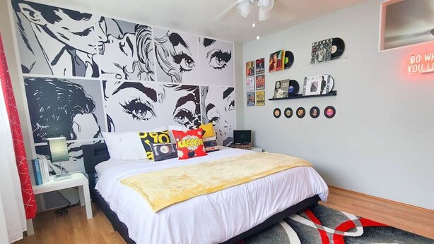 Et soveværelse med en seng, et natbord, en lampe, en væg med illustrationer og en reol med plader.