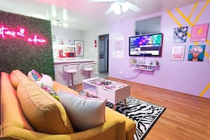 Ein Wohnzimmer mit einer Couch, einem Fernseher, einem Neonzeichen und einer Wand mit Bildern.