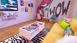 En moderne stue med et fladskærms-tv, et sofabord med magasiner, en gul sofa med farverige puder, og en væg med et stort grafisk tryk.