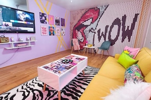 Ein modernes Wohnzimmer mit einem Flachbildfernseher, einem Couchtisch mit Zeitschriften, einem gelben Sofa mit bunten Kissen und einer Wand mit einer großen Grafik.