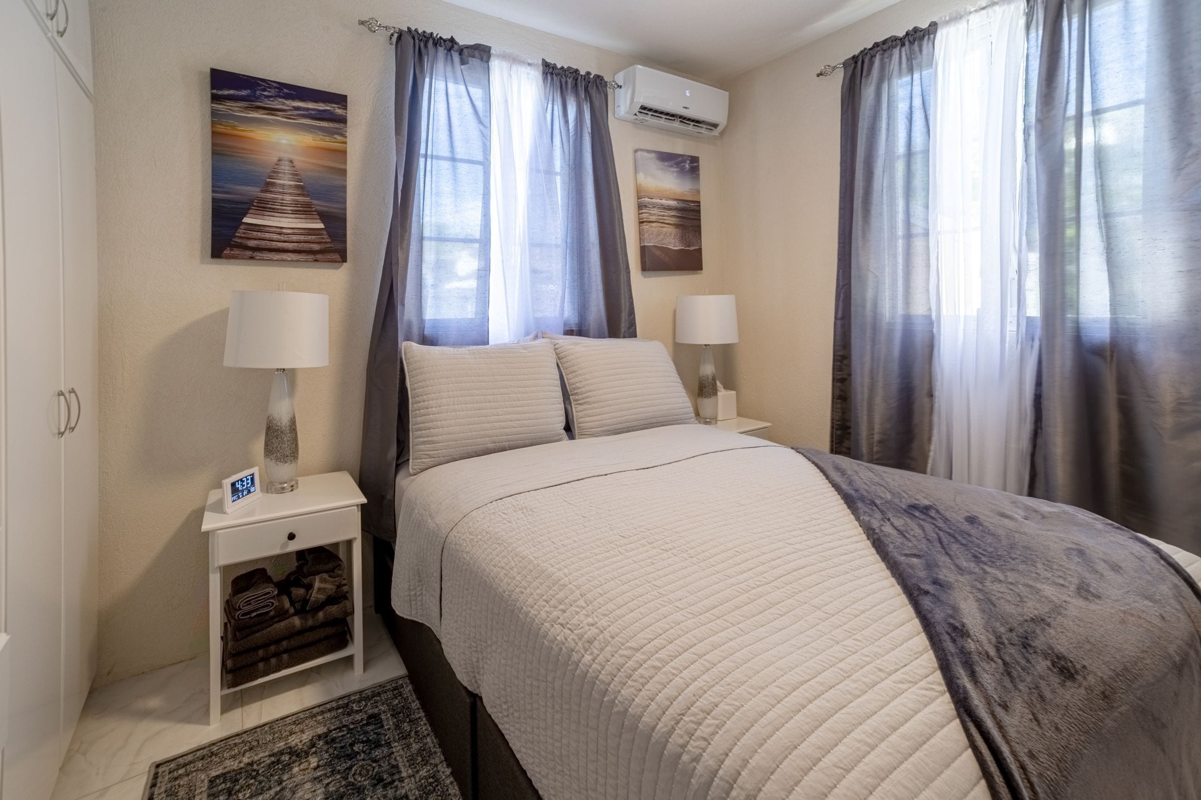 A bedroom with a bed, two bedside tables with lamps, a wall clock, and two paintings.