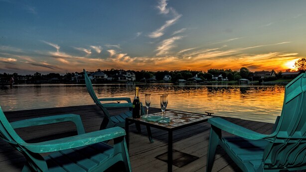 Vista del atardecer desde un muelle con sillas y una mesa, con vista a un lago tranquilo y casas a lo lejos.