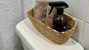 Een badkamer met een wit toilet, een geweven mand met een fles vloeibare zeep en een bruine handdoek, en een badgordijn met een witte textuur.