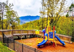 Ein Spielplatz mit Rutsche und Klettergerüsten, umgeben von einem Holzzaun und Bäumen, mit Bergen im Hintergrund.