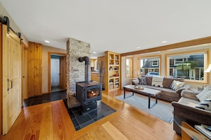 A living room with a stone fireplace, wooden flooring, a sofa, and a view through large windows.