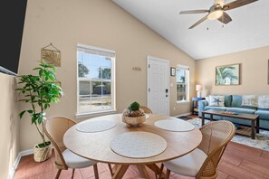 A dining area with a round table, chairs, and a central plant. There is a sofa and a ceiling fan in the background. - New! • Sunset Palms • 2BR • 5 Min Walk to Beach! (Panama City Beach)
