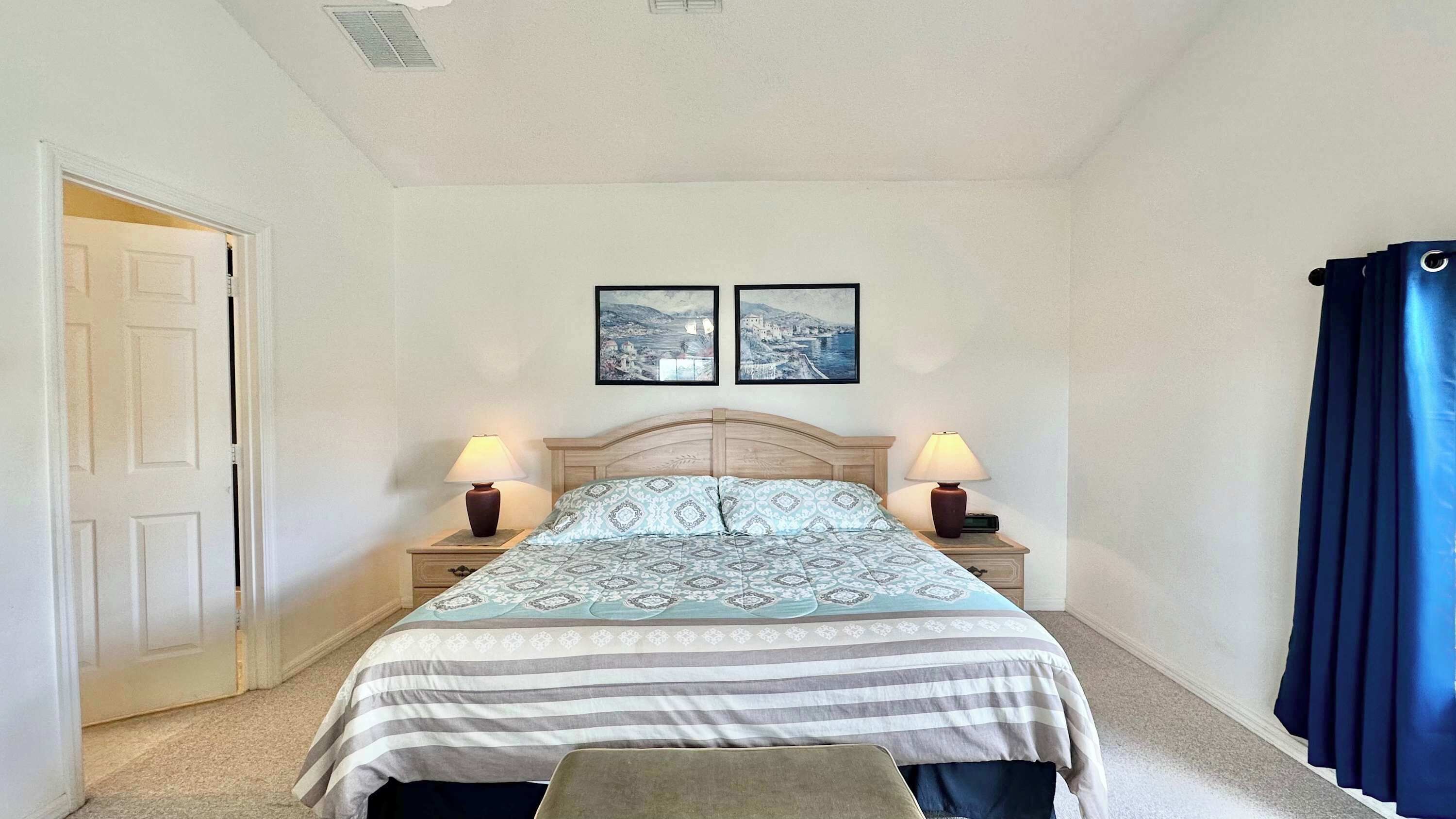 A bedroom with a bed, two bedside tables, a nightstand, a door, and two wall-mounted pictures.