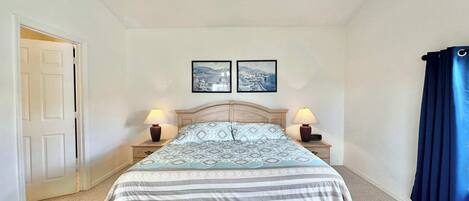 A bedroom with a bed, two bedside tables, a nightstand, a door, and two wall-mounted pictures.