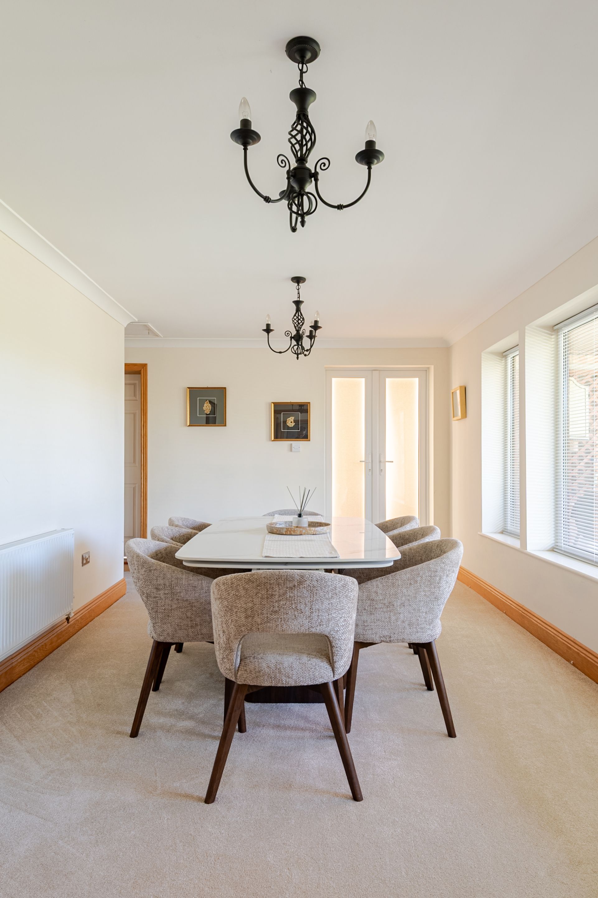 A dining room with a white table, four upholstered chairs, a black chandelier, and framed pictures on the walls.