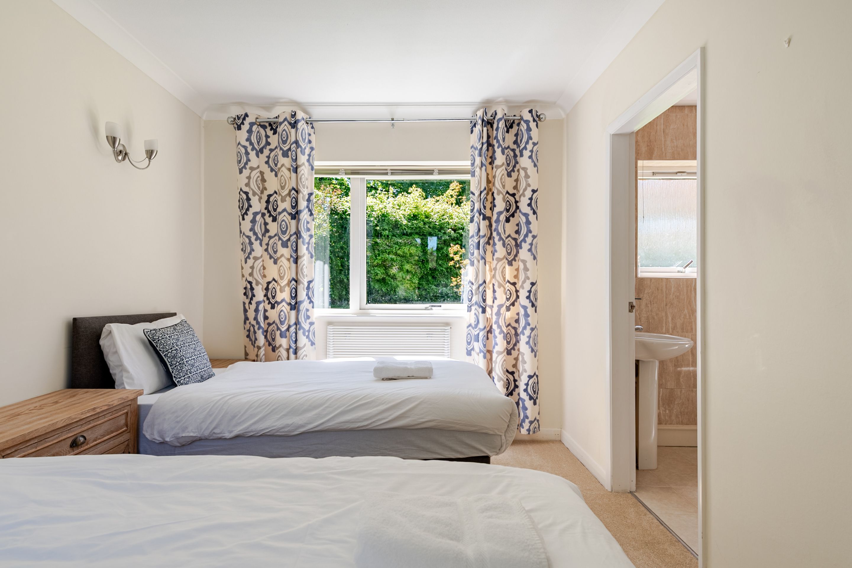 A bedroom with two beds, a window with curtains, a bedside table, and a view of a garden.