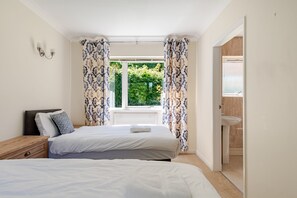 A bedroom with two beds, a window with curtains, a bedside table, and a view of a garden.