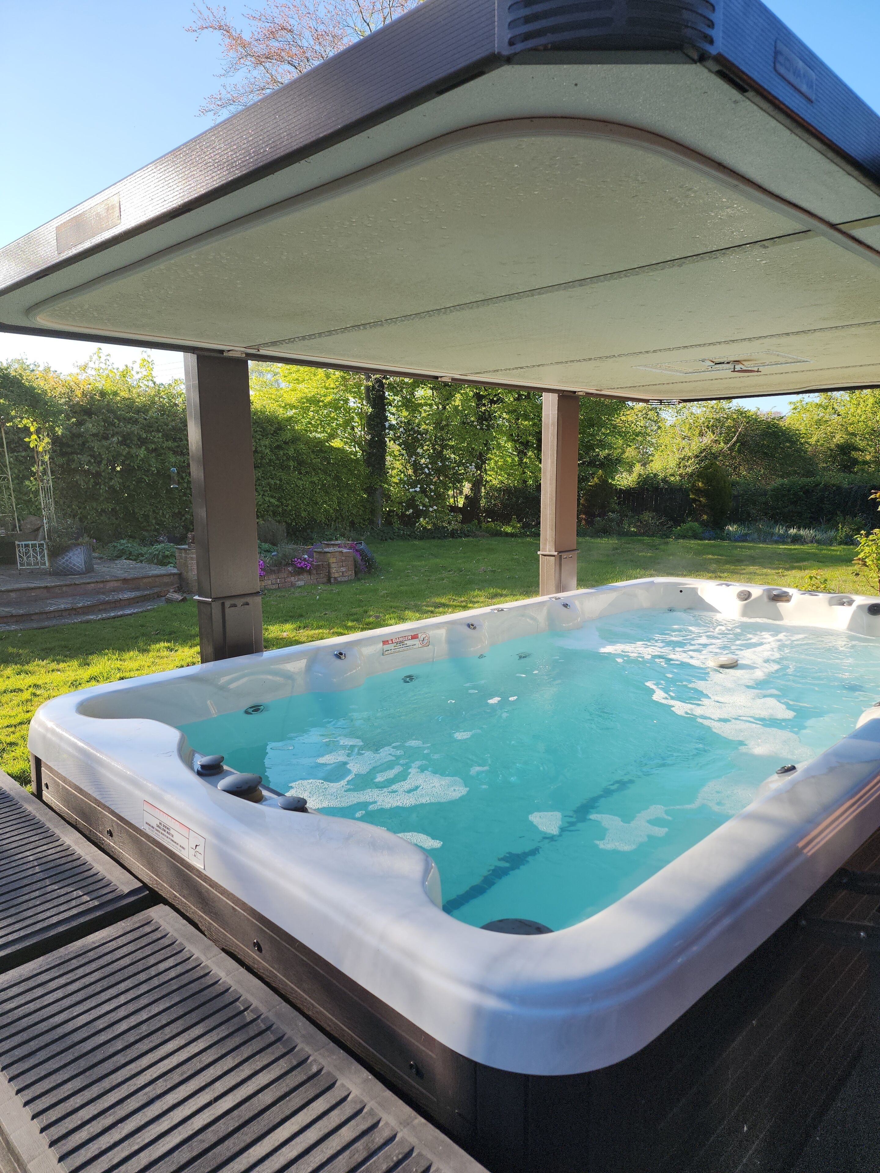 Un jacuzzi bajo un porche cubierto, con un jardín césped y árboles al fondo.