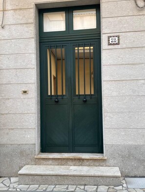A dark green double door with vertical bars, a small window above, and a house number '28' on the wall. - The House Of Egizia (Avola)