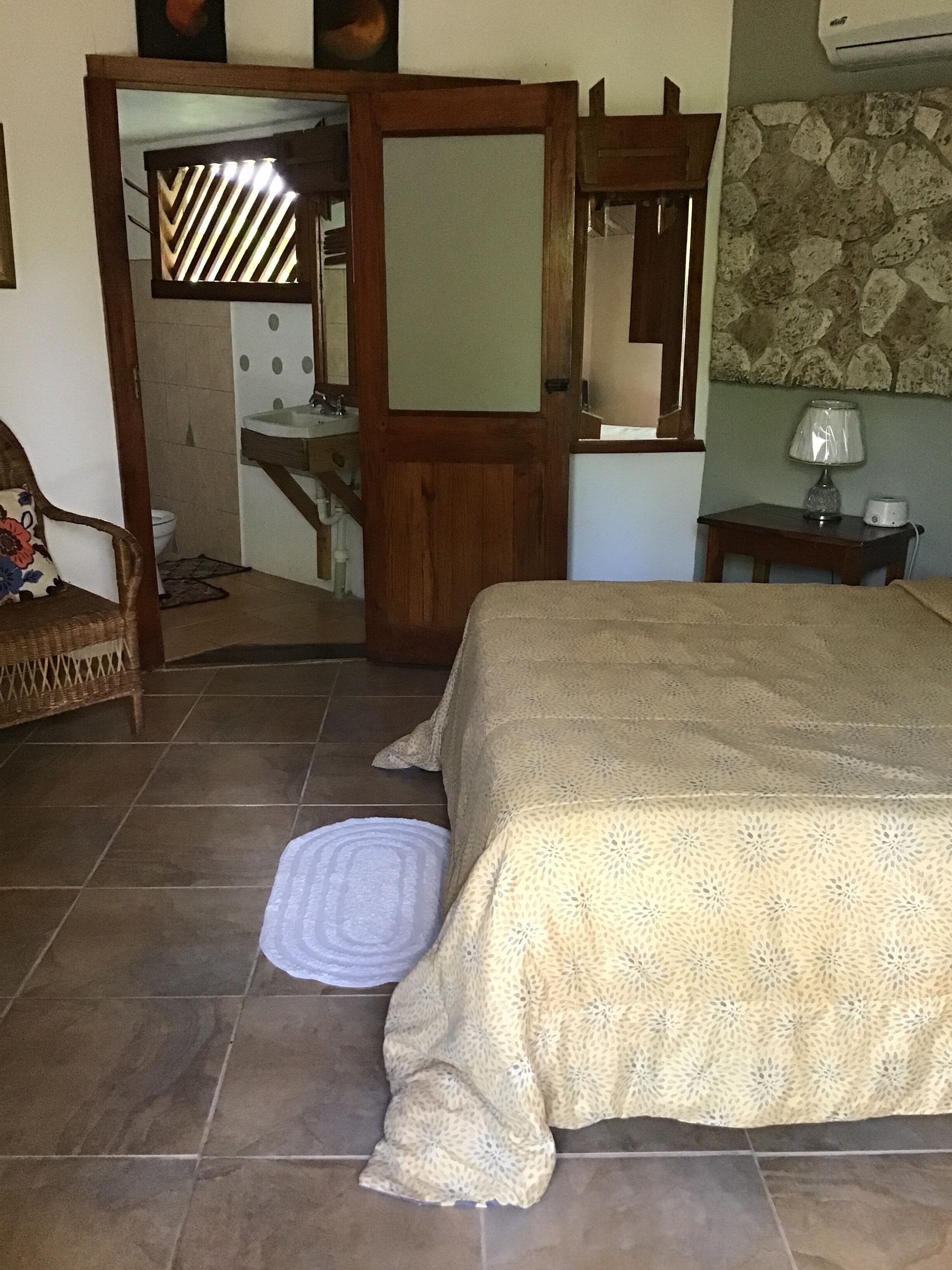 Un dormitorio con cama, una silla, una mesita de noche, una lámpara y un baño visible a través de una puerta abierta.