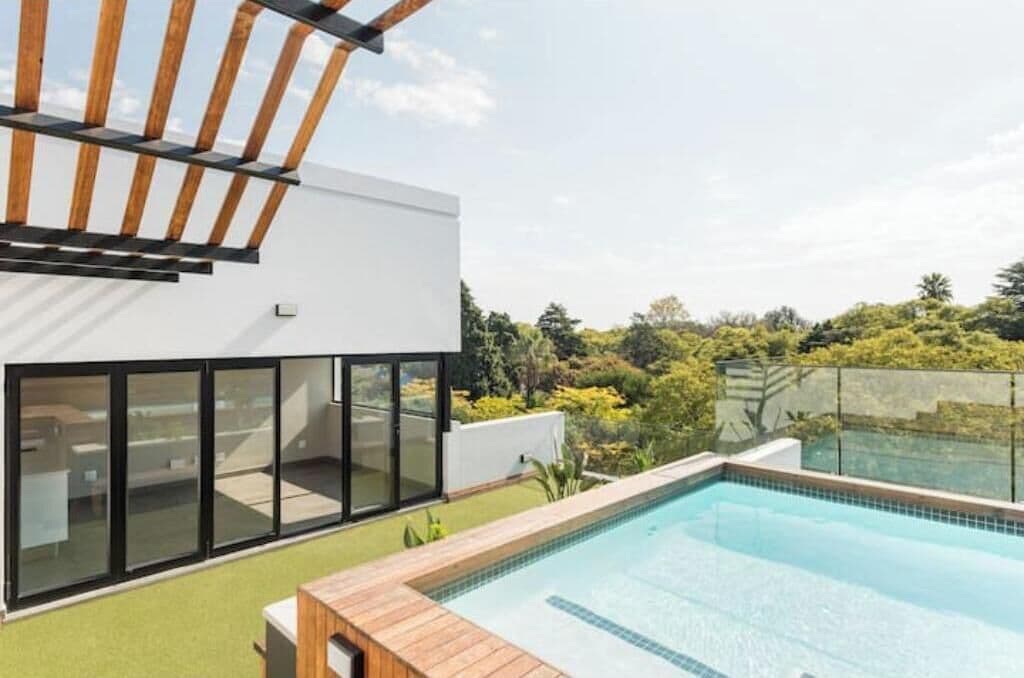 Un edificio moderno con piscina, amplios ventanales de vidrio y una pérgola de madera.