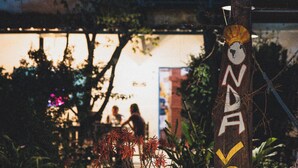 A wooden pole with graffiti, surrounded by vegetation and a building in the background.