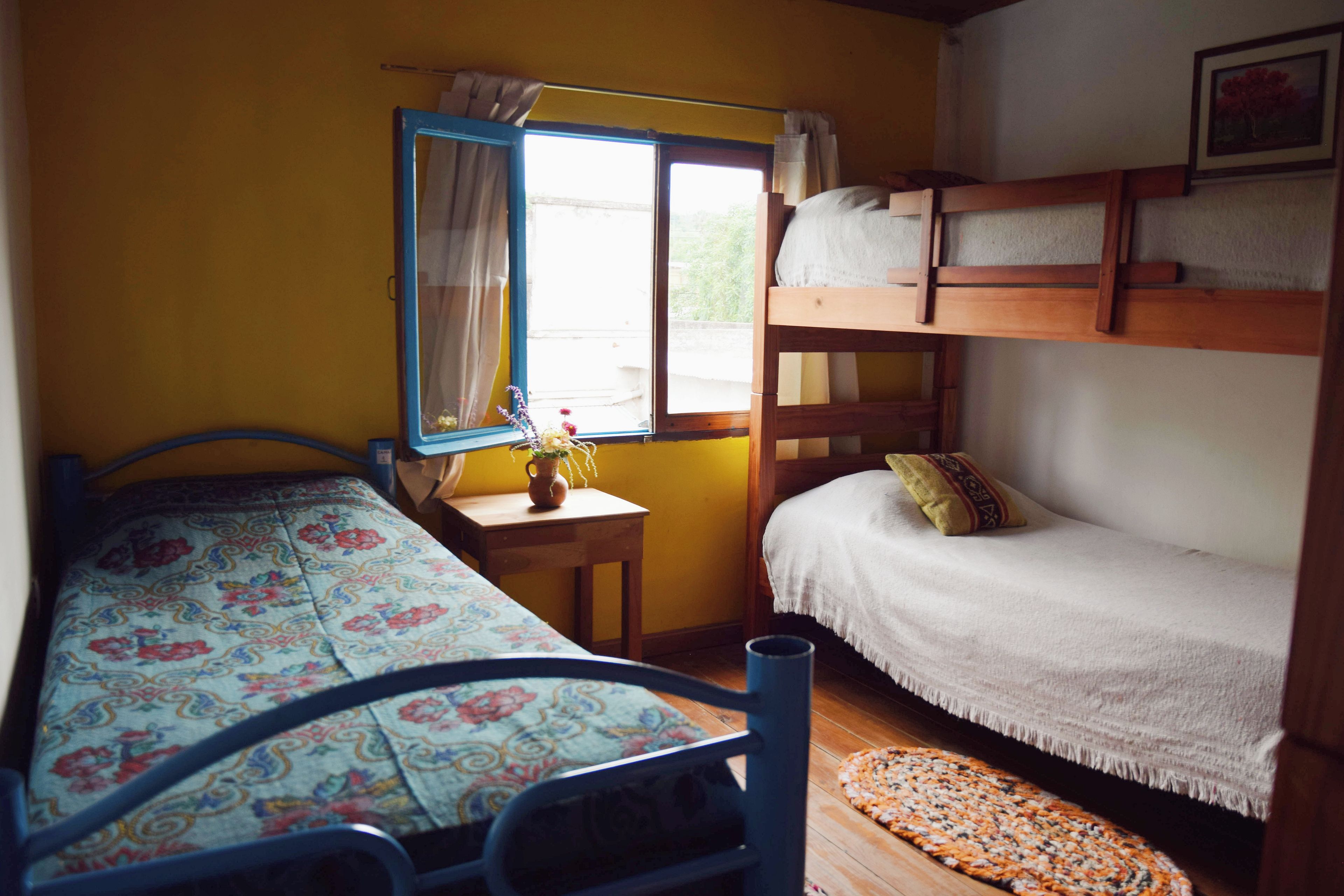 A room with a bunk bed, a single bed, a small table with a vase, and a window with curtains.