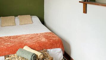 A bedroom with a bed, pillows, a woven chair, a small shelf, a wall hanging, and a mirror.