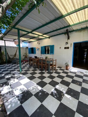 A covered patio with a checkered floor, wooden tables and chairs, a hammock, and a potted plant.