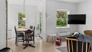 A modern living room with a dining area, a TV mounted on the wall, and a view of greenery through the windows. - Cozy cottage in Nykøbing Sjælland (Nykøbing Sj)
