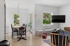 A modern living room with a dining area, a TV mounted on the wall, and a view of greenery through the windows. - Cozy cottage in Nykøbing Sjælland (Nykøbing Sj)