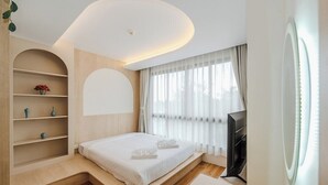 A modern bedroom with a bed, built-in shelving, a flat-screen TV, and a large window with curtains. - Sunset View Condo Steps from Karon Beach #C162 (Tambon Karon)