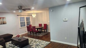 A living room with a dining area, a ceiling fan, a chandelier, and a window with blinds.