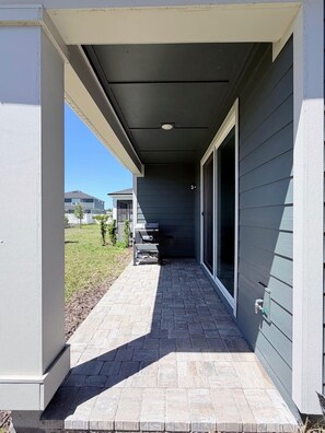 Ein überdachter Patio-Bereich mit Grill und Gartenmöbeln, der zu einer Glastür mit Schiebeöffnung führt.