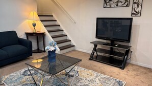A living room with a sofa, a coffee table, a TV on a stand, a lamp, and a staircase.