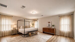 Een slaapkamer met een hemelbed, een nachtkastje met een lamp, een houten dressoir en een groot raam met gordijnen.