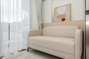 Ein beiges Sofa in einem Zimmer mit weißem Boden, einem Fenster mit transparenten Vorhängen und einem gerahmten abstrakten Kunstwerk an der Wand.