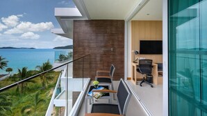 A modern balcony with a view of the sea, a dining area with chairs and a table, and a room with a TV and desk.