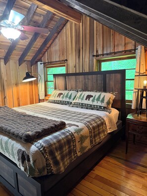 Ein schlicht eingerichtetes Schlafzimmer im Holzblockhaus-Stil mit Bett, Nachttisch, Lampe und Fenster mit Blick auf grüne Landschaft.