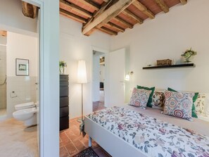 A bedroom with a bed, a bedside table, a lamp, a shelf with decorative items, and a bathroom with a toilet and sink. - Apartment in Tuscany With Stunning Views (Capannori)