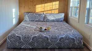 A bedroom with a wooden ceiling, a patterned bedspread, a bedside table with a basket, and a window. - Freedom-Cabin-1 @ Eagle Hollow (Goreville)
