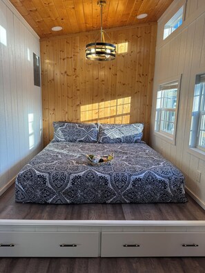 Ein Schlafzimmer mit Holzboden, einer gemusterten Bettdecke, einem Nachttisch mit Korb und einem Fenster.