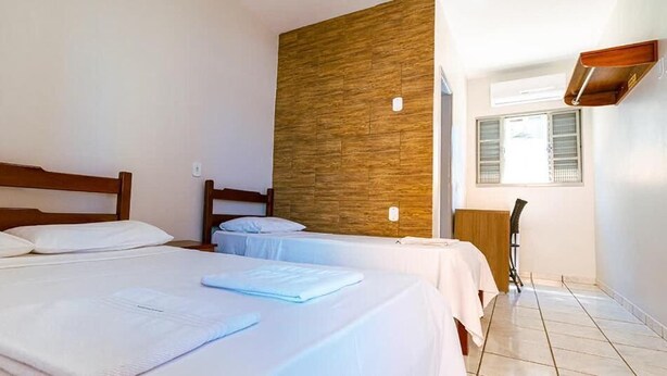 A hotel room with two beds, a wooden accent wall, a desk with a chair, and a window with blinds.