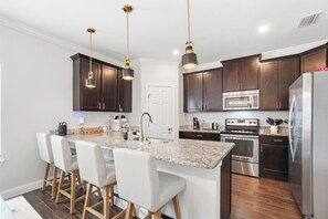 A modern kitchen with dark wood cabinets, stainless steel appliances, a granite countertop, and bar stools. - Luxe Beach Retreat | Pool • Arcade • Walk to Beach (Jacksonville Beach)