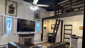 Una sala de estar con chimenea, un televisor de pantalla plana grande, un sofá de cuero, un ventilador de techo y una cocina con una escalera.